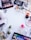 A collection of various makeup and skincare products arranged on a white surface. The items include eyeshadow palettes, false eyelashes, brushes, lipsticks, foundation, eyelash curler, and other cosmetics. There are also decorative white flowers scattered around the products.