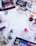 A collection of various makeup and skincare products arranged on a white surface. The items include eyeshadow palettes, false eyelashes, brushes, lipsticks, foundation, eyelash curler, and other cosmetics. There are also decorative white flowers scattered around the products.