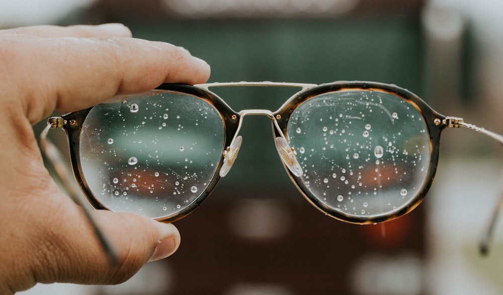 eine Hand, die eine Brille mit Wassertropfen darauf hält