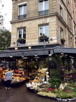 A charming flower shop located in a corner building, featuring an array of vibrant flowers displayed outside. The shop's awning reads 'O' Fleurs de Montmartre,' surrounded by lush green plants and floral arrangements. A few people are browsing the floral selections under the soft lighting that contributes to a quaint and welcoming atmosphere.