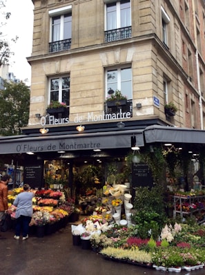 A charming flower shop located in a corner building, featuring an array of vibrant flowers displayed outside. The shop's awning reads 'O' Fleurs de Montmartre,' surrounded by lush green plants and floral arrangements. A few people are browsing the floral selections under the soft lighting that contributes to a quaint and welcoming atmosphere.