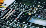 A close-up of a DJ mixing console with various knobs, sliders, buttons, and LED lights. The console is branded and features multiple channels, effects, and controls for music production or live DJing.