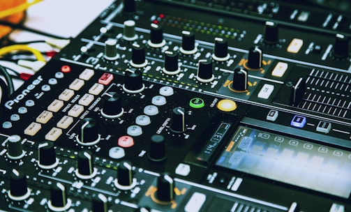 A close-up of a DJ mixing console with various knobs, sliders, buttons, and LED lights. The console is branded and features multiple channels, effects, and controls for music production or live DJing.