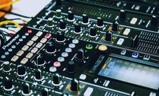 A close-up of a DJ mixing console with various knobs, sliders, buttons, and LED lights. The console is branded and features multiple channels, effects, and controls for music production or live DJing.