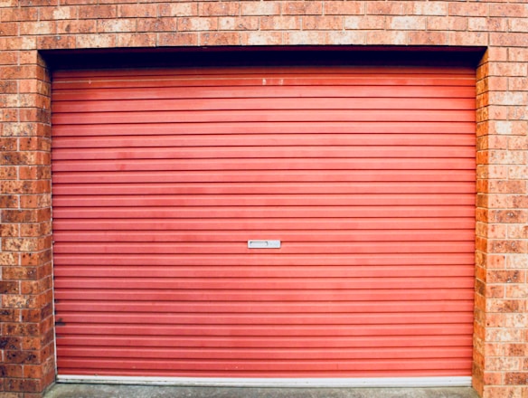 A closed red garage door with horizontal panels is set within a brick wall. The bricks are red with various shades, showing some texture and detail.