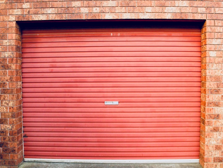 A closed red garage door with horizontal panels is set within a brick wall. The bricks are red with various shades, showing some texture and detail.