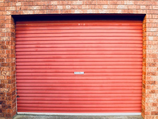 A closed red garage door with horizontal panels is set within a brick wall. The bricks are red with various shades, showing some texture and detail.