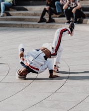 A person is breakdancing on a paved area, balancing on one hand with their legs bent in the air, wearing a white tracksuit with red and blue stripes. A few people are sitting on steps in the background, casually observing the performance.