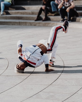 A person is breakdancing on a paved area, balancing on one hand with their legs bent in the air, wearing a white tracksuit with red and blue stripes. A few people are sitting on steps in the background, casually observing the performance.