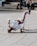 A person is breakdancing on a paved area, balancing on one hand with their legs bent in the air, wearing a white tracksuit with red and blue stripes. A few people are sitting on steps in the background, casually observing the performance.