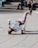 A person is breakdancing on a paved area, balancing on one hand with their legs bent in the air, wearing a white tracksuit with red and blue stripes. A few people are sitting on steps in the background, casually observing the performance.