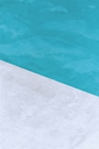 A close-up view of a pool with clear blue water and a corner of light gray stone or concrete coping. The contrast between the water and the poolside creates a clean, refreshing look.