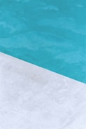 A close-up view of a pool with clear blue water and a corner of light gray stone or concrete coping. The contrast between the water and the poolside creates a clean, refreshing look.
