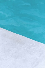 A close-up view of a pool with clear blue water and a corner of light gray stone or concrete coping. The contrast between the water and the poolside creates a clean, refreshing look.