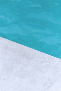 A close-up view of a pool with clear blue water and a corner of light gray stone or concrete coping. The contrast between the water and the poolside creates a clean, refreshing look.