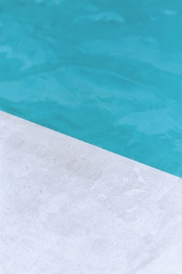 A close-up view of a pool with clear blue water and a corner of light gray stone or concrete coping. The contrast between the water and the poolside creates a clean, refreshing look.