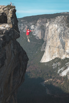 A person in a red shirt is balancing on a slackline stretched high above a canyon. The scene is set against a backdrop of towering cliffs and a dense forest. The sky is clear and blue, adding a serene contrast to the daring act.