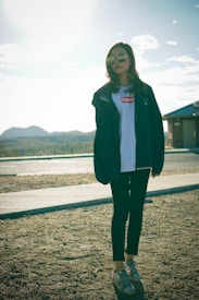 A person wearing sunglasses and casual clothing, including a white t-shirt with a prominent logo, a dark jacket, and jeans, is standing on a sunlit, gravelly surface. The background features distant hills under a partly cloudy sky.
