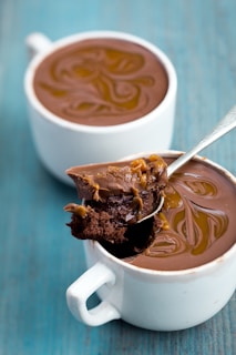 Two white mugs containing a rich, chocolate dessert topped with swirled caramel. A spoon is lifting a portion from one of the mugs, revealing a moist, dense texture underneath.