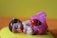 A small figurine of a sleeping baby with a pacifier in its mouth, dressed in a pink cloth with a colorful ladybug decoration. The baby has brown hair with a purple decoration on top. The figurine is set against a bright yellow and orange background.