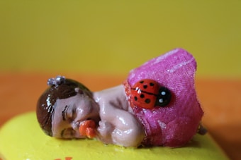 A small figurine of a sleeping baby with a pacifier in its mouth, dressed in a pink cloth with a colorful ladybug decoration. The baby has brown hair with a purple decoration on top. The figurine is set against a bright yellow and orange background.