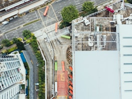 Aerial view of an industrial area with a large warehouse on the right side, multiple shipping containers, and trucks parked in an organized manner. The adjacent road shows traffic markings, a single red bus, and several cars. Vegetation and buildings are visible, adding an urban feel.