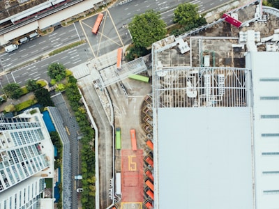 Aerial view of an industrial area with a large warehouse on the right side, multiple shipping containers, and trucks parked in an organized manner. The adjacent road shows traffic markings, a single red bus, and several cars. Vegetation and buildings are visible, adding an urban feel.