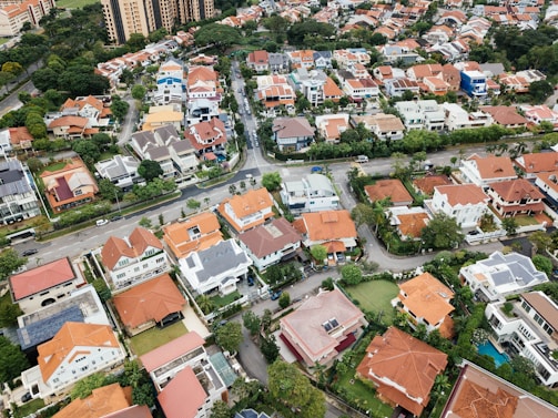 Aerial view of a suburban neighborhood featuring various houses with red-tiled and gray roofs, neatly arranged along curved roads and surrounded by green vegetation. Tall apartment buildings and dense trees are visible in the background.