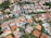 Aerial view of a suburban neighborhood featuring various houses with red-tiled and gray roofs, neatly arranged along curved roads and surrounded by green vegetation. Tall apartment buildings and dense trees are visible in the background.