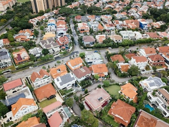 Aerial view of a suburban neighborhood featuring various houses with red-tiled and gray roofs, neatly arranged along curved roads and surrounded by green vegetation. Tall apartment buildings and dense trees are visible in the background.