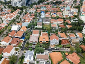 Aerial view of a residential neighborhood featuring a collection of closely packed houses with predominantly orange roofs. The area includes neatly arranged streets and green spaces around the buildings, giving an orderly appearance. In the background, larger multi-story buildings can be seen, indicating an urban setting.