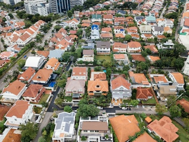 Aerial view of a residential neighborhood featuring a collection of closely packed houses with predominantly orange roofs. The area includes neatly arranged streets and green spaces around the buildings, giving an orderly appearance. In the background, larger multi-story buildings can be seen, indicating an urban setting.