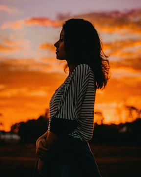 A silhouette of a person with long hair appears against a dramatic sunset. The sky is filled with vibrant orange and red hues, with fluffy clouds adding texture. The person is facing sideways, wearing a striped shirt.