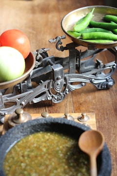 A rustic scene featuring an antique balance scale with brass pans, containing a tomato and an apple on one side and several green chili peppers on the other. Below the scale is a stone bowl filled with a green mixture, accompanied by a wooden spoon and a set of metal weights.