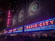 Neon signs with bright red letters spell out 'Radio City' on the facade of a large building, illuminated in vibrant colors. Blues and reds dominate the signage, with decorative emblems visible on the upper portion of the building. The scene is bustling with people lined up along barriers, suggesting an event or a show is upcoming.