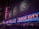 Neon signs with bright red letters spell out 'Radio City' on the facade of a large building, illuminated in vibrant colors. Blues and reds dominate the signage, with decorative emblems visible on the upper portion of the building. The scene is bustling with people lined up along barriers, suggesting an event or a show is upcoming.
