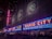 Neon signs with bright red letters spell out 'Radio City' on the facade of a large building, illuminated in vibrant colors. Blues and reds dominate the signage, with decorative emblems visible on the upper portion of the building. The scene is bustling with people lined up along barriers, suggesting an event or a show is upcoming.