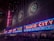 Neon signs with bright red letters spell out 'Radio City' on the facade of a large building, illuminated in vibrant colors. Blues and reds dominate the signage, with decorative emblems visible on the upper portion of the building. The scene is bustling with people lined up along barriers, suggesting an event or a show is upcoming.