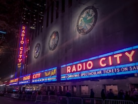 Neon signs with bright red letters spell out 'Radio City' on the facade of a large building, illuminated in vibrant colors. Blues and reds dominate the signage, with decorative emblems visible on the upper portion of the building. The scene is bustling with people lined up along barriers, suggesting an event or a show is upcoming.