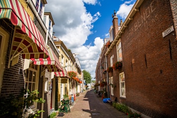 A picturesque narrow street lined with traditional brick buildings adorned with flower boxes and striped awnings. The bright blue sky with scattered fluffy clouds complements the vibrant flowers and adds a cheerful atmosphere. The street is clean and inviting, with a small hotel sign visible.