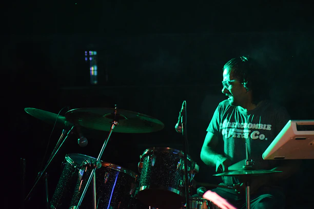 A person wearing glasses and a branded t-shirt is playing a drum set under dim lighting conditions. The scene suggests a live performance or practice session. The drum set includes cymbals and other percussion instruments, surrounded by a dark background.
