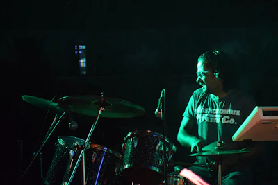 A person wearing a stylish drum-themed t-shirt, standing by a drum kit with colorful lighting.