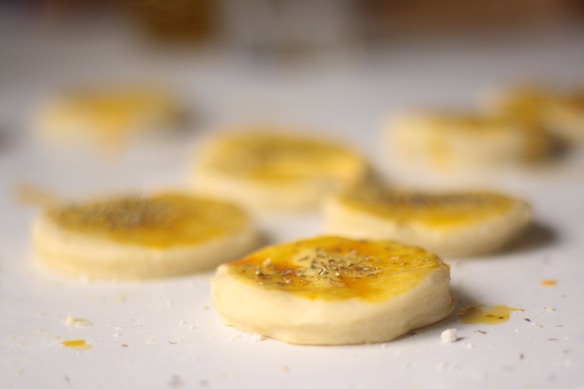 A close-up view of raw dough slices on a surface, each topped with a golden glaze and sprinkled with herbs. The focus is on the nearest slice, with others blurred in the background.