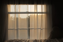 Soft sunlight filters through sheer curtains, casting a warm, gentle glow into the room. The outside scenery is slightly blurred, enhancing the calm and serene atmosphere. A hint of a bed with wrinkled sheets is visible, adding a cozy and tranquil vibe.