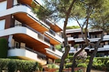 A modern apartment building with distinctively rounded balconies is partially obscured by lush green trees. The architecture prominently features wooden and white elements, and neat hedges border the building, enhancing the aesthetic with well-maintained greenery. Several large windows and sliding doors indicate access to outdoor living spaces.