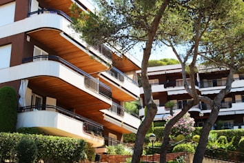A modern apartment building with distinctively rounded balconies is partially obscured by lush green trees. The architecture prominently features wooden and white elements, and neat hedges border the building, enhancing the aesthetic with well-maintained greenery. Several large windows and sliding doors indicate access to outdoor living spaces.