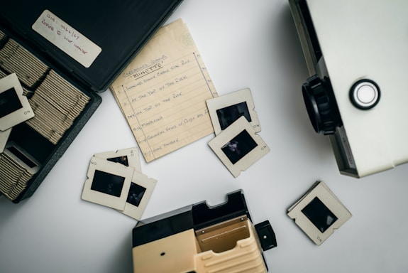 A collection of vintage photographic slides and a projector are displayed on a flat surface. The slides are housed in a black case, with several scattered around, revealing film strips. A handwritten note, possibly a list of slide subjects or order, lies amidst the slides.
