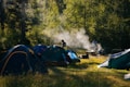 A campsite is set in a lush forest with several tents arranged on a grassy area. Wisps of smoke rise from a small campfire, suggesting cooking or warmth. The sunlight filters through the trees, casting dappled shadows on the ground. Two figures are present, one near the tents and another by the fire, enhancing the sense of outdoor activity and leisure.