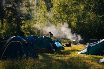 A campsite is set in a lush forest with several tents arranged on a grassy area. Wisps of smoke rise from a small campfire, suggesting cooking or warmth. The sunlight filters through the trees, casting dappled shadows on the ground. Two figures are present, one near the tents and another by the fire, enhancing the sense of outdoor activity and leisure.