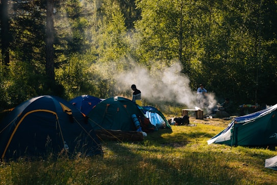 A campsite is set in a lush forest with several tents arranged on a grassy area. Wisps of smoke rise from a small campfire, suggesting cooking or warmth. The sunlight filters through the trees, casting dappled shadows on the ground. Two figures are present, one near the tents and another by the fire, enhancing the sense of outdoor activity and leisure.